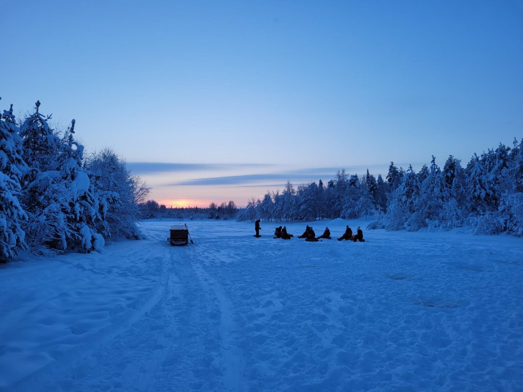 Talvimaisema auringonlaskun aikaan ja ihmisryhmä pilkkimässä lumisessa ympäristössä.
