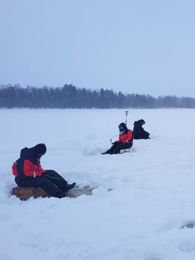 Henkilö pilkkimässä järven jäällä, ympärillä luminen metsä ja harmaa taivas