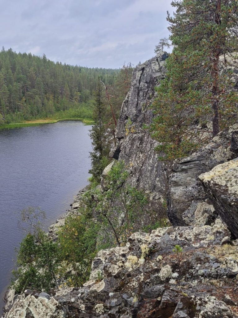 Jyrkkä kallionseinämä, taustalla metsä ja virtaava järvi kesäisessä maisemassa.