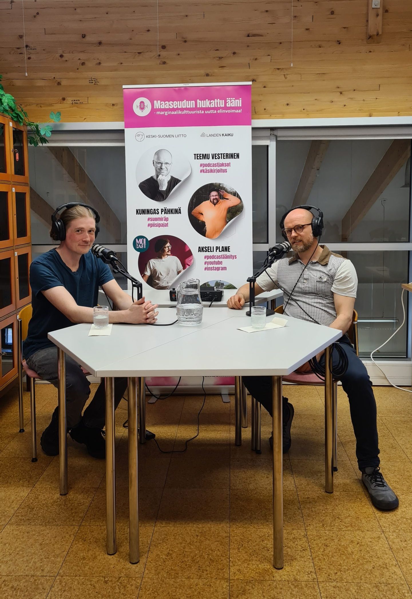 Kaksi miestä istuu podcast-studiossa kuulokkeet korvilla ja katsoo kameraan.