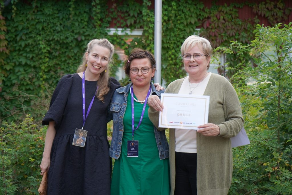 Kolme erikäistä naista seisomassa iloiten diplomi kädessä vehreällä pihalla.