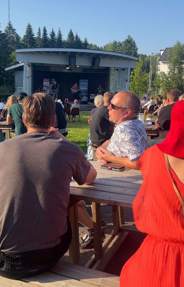 Joukko ihmisiä istumassa helteisessä puistossa, katsomassa keikkaa.