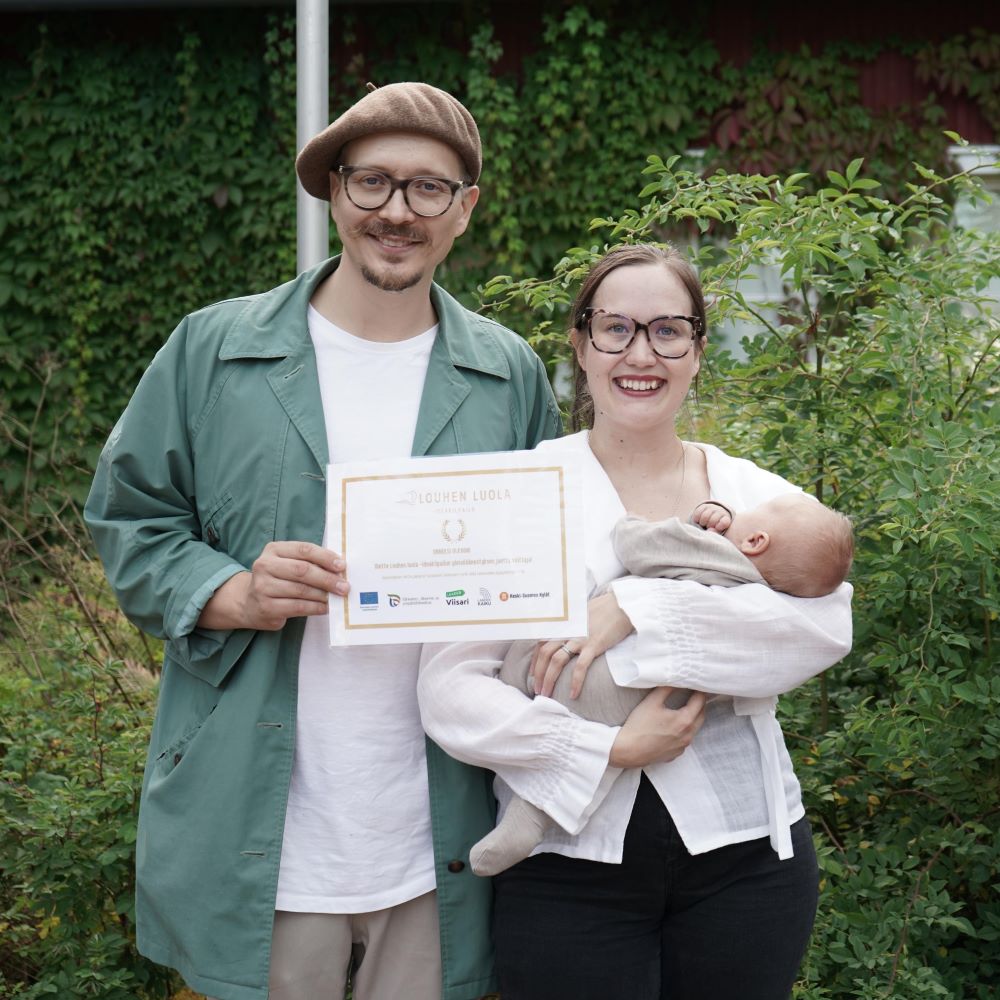 nuori pariskunta ja muutaman kuukaiden ikäinen vauva katsovat kameraan ja pitävät edessään diplimia vehreällä pihamaalla